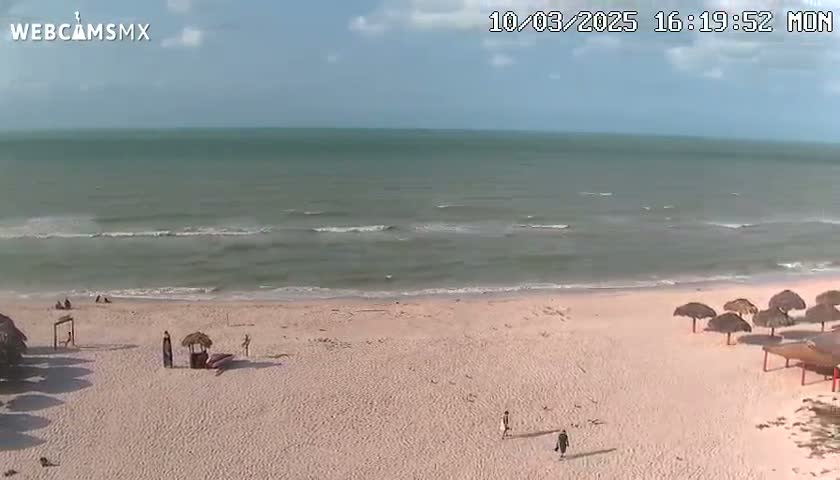 A mostly sunny day at a beach shows calm waves lapping a light-pink sandy shore where a few people are walking and sitting under thatched umbrellas.
