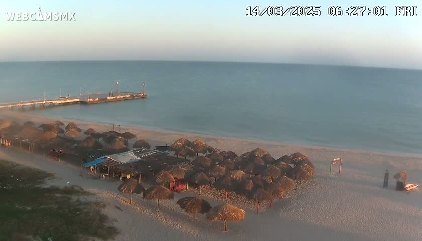 A beach with numerous thatched umbrellas, a pier extending into a calm sea under a clear, dawn sky.