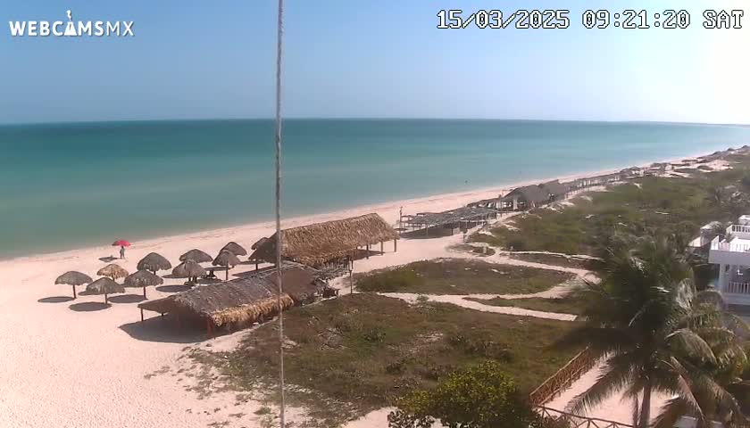 A sandy beach with thatched-roof huts and calm, light-blue water under a clear sky.
