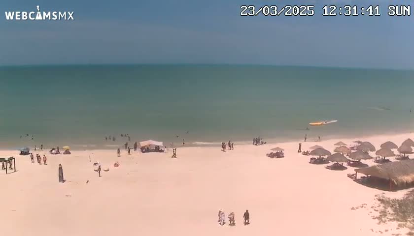 A mostly sunny beach scene shows a calm, light-green ocean, a sandy shore with scattered beachgoers and umbrellas, and a few people near the water's edge.