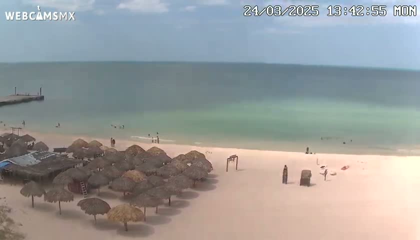 A mostly sunny beach scene shows calm, light-green water, a sandy shore with numerous thatched umbrellas, a small pier, and a few people scattered on the beach and in the water.