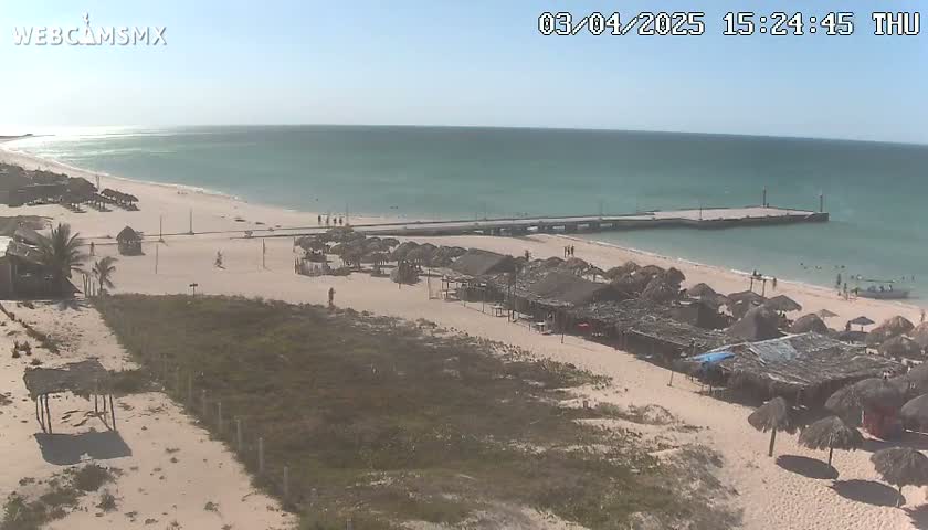 A sandy beach with numerous thatched-roof structures and a long pier extends into a calm, light-blue ocean under a clear, sunny sky.