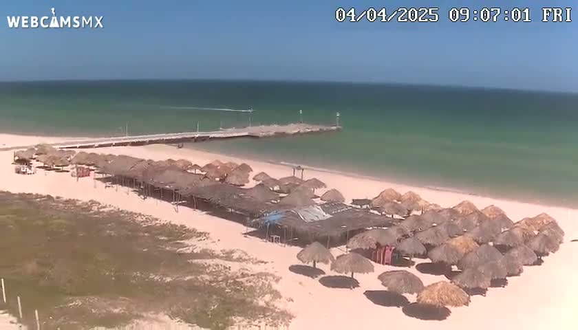 A sandy beach with numerous thatched umbrellas, a pier extending into a calm, clear turquoise ocean under a bright sunny sky.