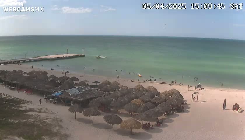 A sandy beach with numerous thatched umbrellas, a pier, and a calm, light-green ocean under a partly sunny sky.