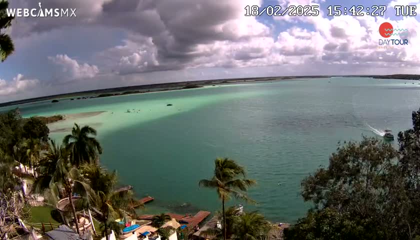 A partly cloudy sky overlooks a tropical lagoon with calm, teal-green water, palm trees, and a few boats.
