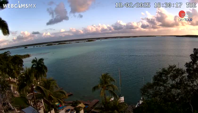 A tranquil body of water, under a partly cloudy sunset sky, is surrounded by lush green vegetation and a few boats are visible in the distance.