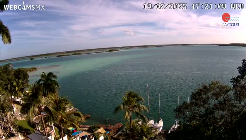 An expansive body of teal-colored water, partially enclosed by land, is viewed from an elevated vantage point on a partly sunny day; several sailboats are moored near the shoreline, which is lined with lush tropical vegetation.