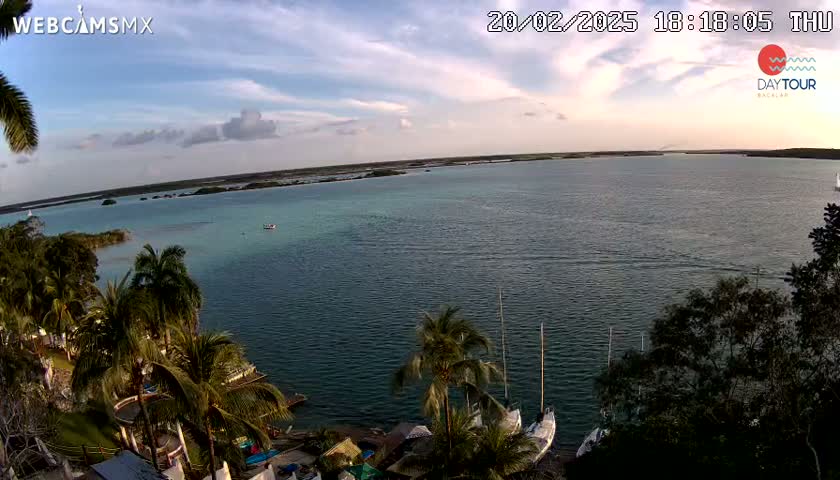 An expansive body of calm, teal water is seen from a high vantage point, under a partly cloudy sky at sunset, with palm trees and sailboats visible along the shoreline.