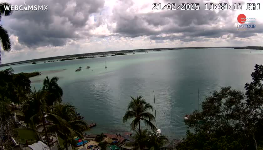 A partly cloudy day overlooking a calm teal bay with several boats and palm trees lining the shore.