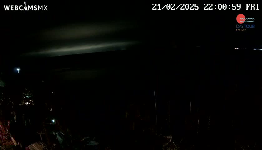 A nighttime, dark view of a body of water, possibly a lake or bay, with dimly lit land visible in the foreground under a mostly cloudy sky.