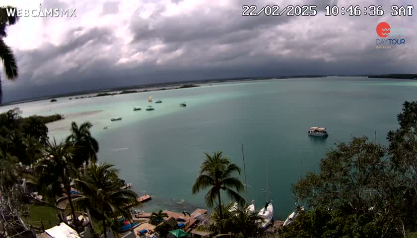 A tranquil teal lagoon under a cloudy sky is dotted with several small boats and sailboats, viewed from a high vantage point overlooking lush tropical vegetation.