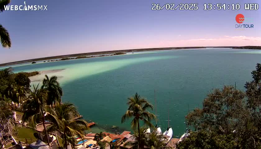 A tranquil, teal-colored lagoon is seen from an elevated viewpoint on a sunny day, with palm trees and a few sailboats visible near the shore.