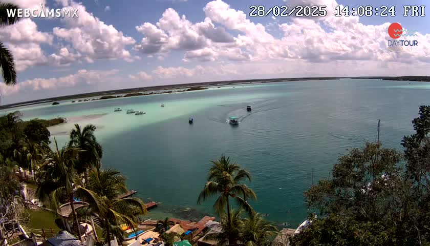 A partly cloudy day reveals a tranquil turquoise bay dotted with several small boats, viewed from an elevated vantage point overlooking a lush, tropical shoreline.