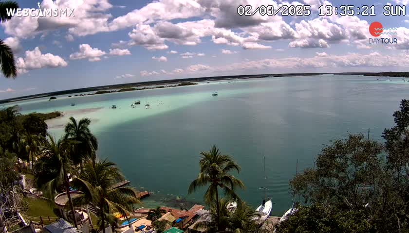A partly cloudy sunny day overlooks a calm teal-green body of water with several boats, palm trees, and buildings along the shoreline.