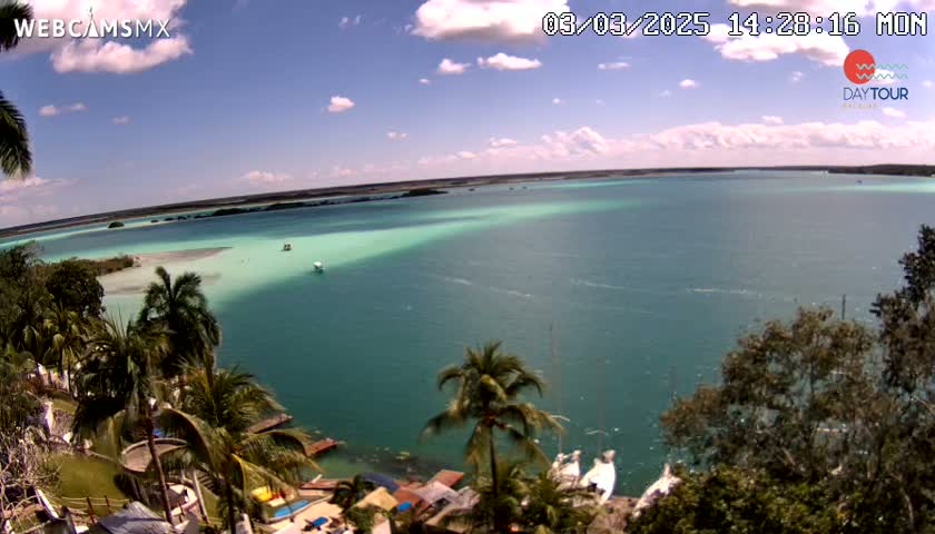 A panoramic view shows a partly cloudy sky over a calm, teal-colored body of water, lined with tropical vegetation and a few boats.