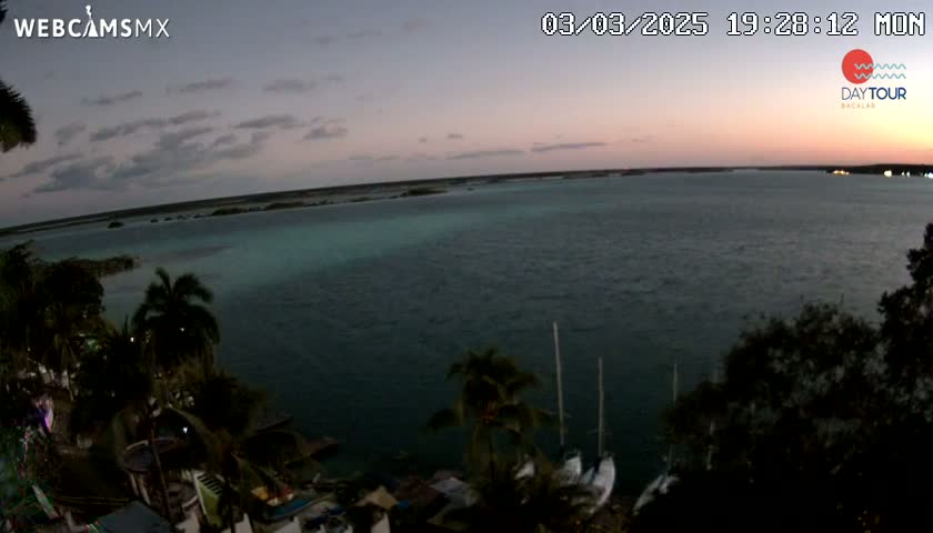 An expansive body of dark, calm water is seen at dusk, under a mostly clear sky, with tropical vegetation and a few sailboats visible along the shoreline.
