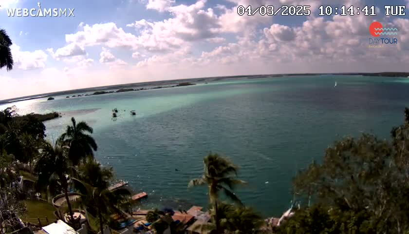 A partly cloudy day reveals a tranquil turquoise body of water, dotted with small boats and surrounded by lush green vegetation.