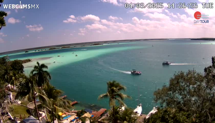 An aerial view shows a tropical lagoon with turquoise water, several boats, and palm trees lining a shoreline under a partly cloudy sky.