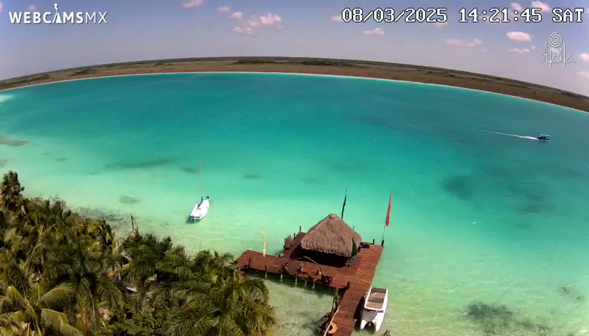 An aerial view shows a vibrant turquoise lagoon under a partly sunny sky, with a small thatched-roof structure on a wooden dock, several boats on the water, and lush tropical vegetation along the shore.