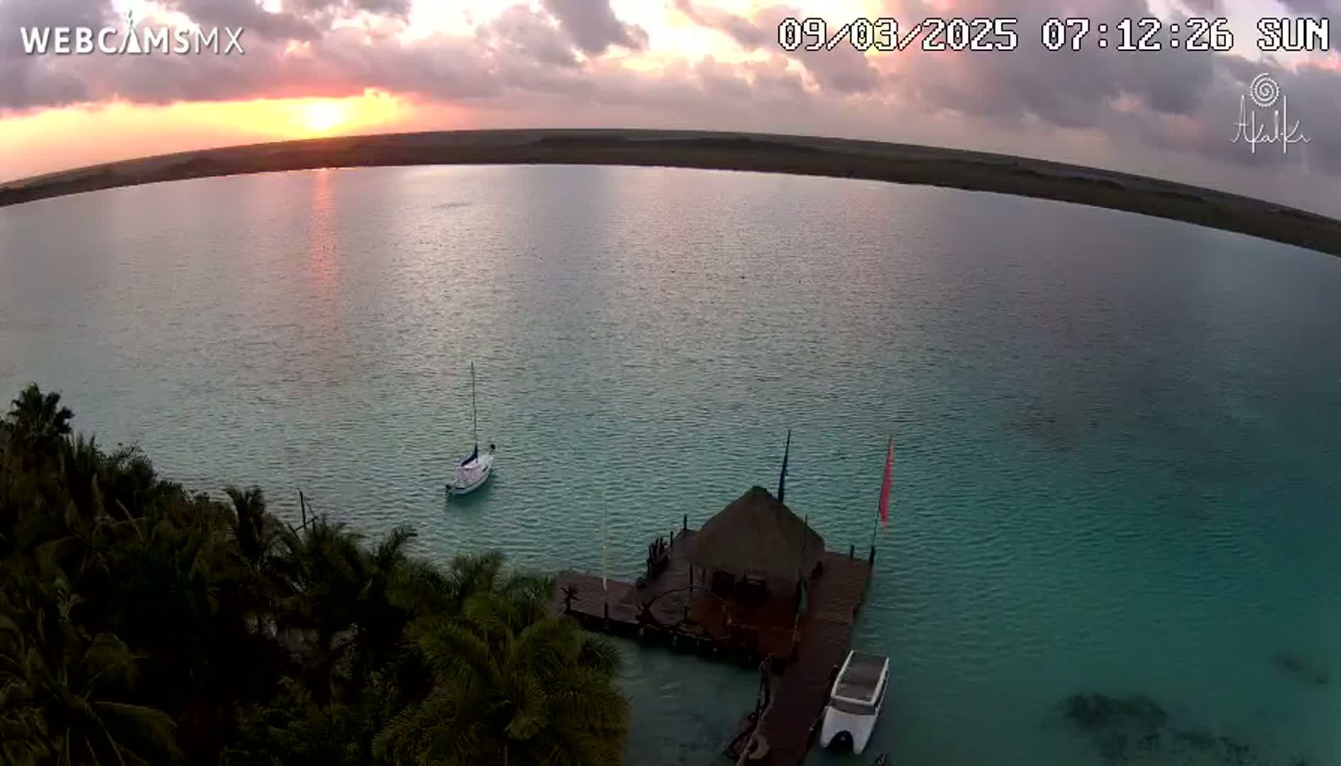 An aerial view shows a calm, teal body of water at sunrise, with a small sailboat and a dock with a thatched-roof structure and a small boat moored nearby, all surrounded by lush tropical vegetation.