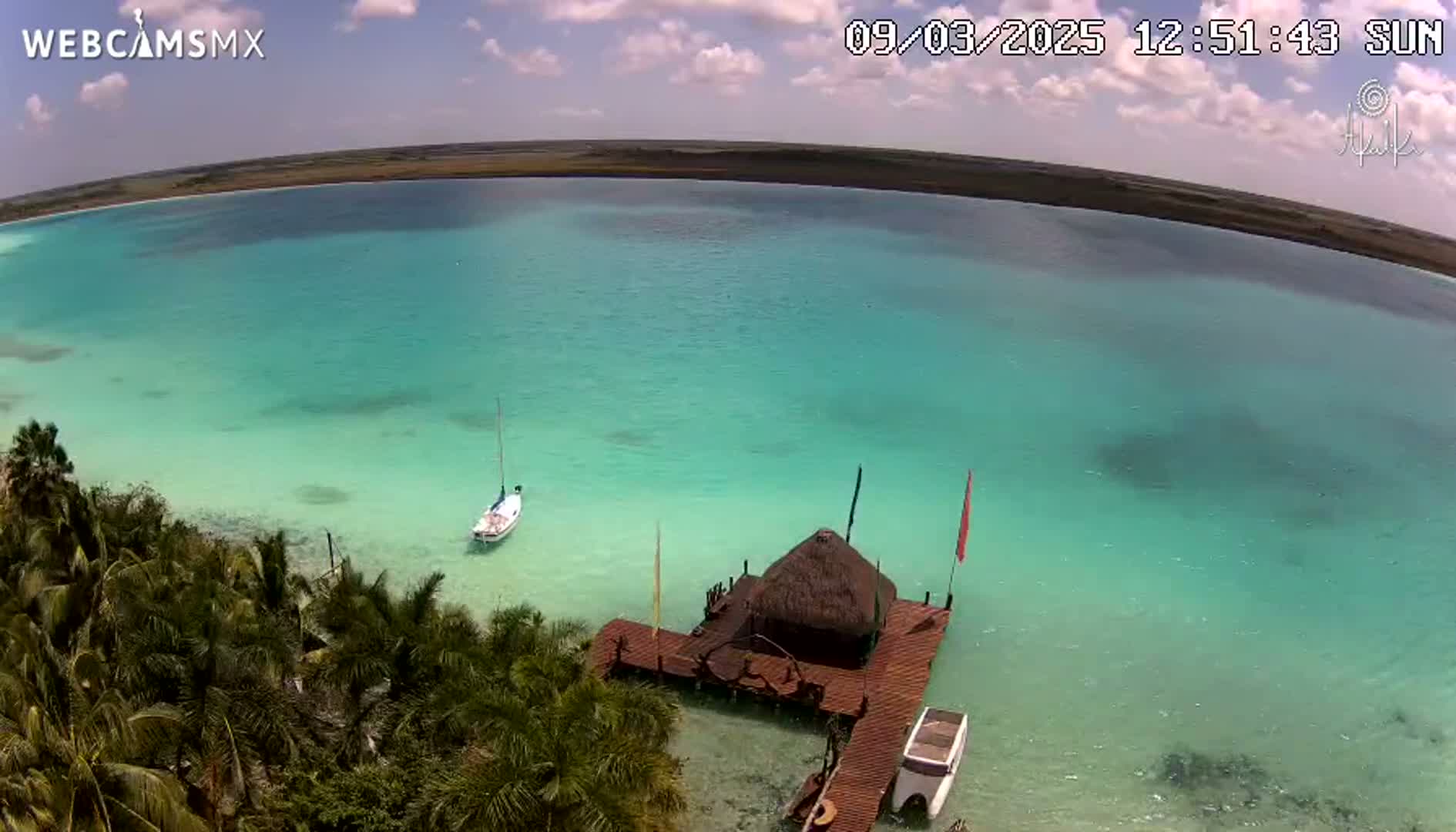 An aerial view shows a calm, turquoise body of water with a small sailboat and a thatched-roof structure on a dock, surrounded by lush green vegetation under a partly cloudy sky.