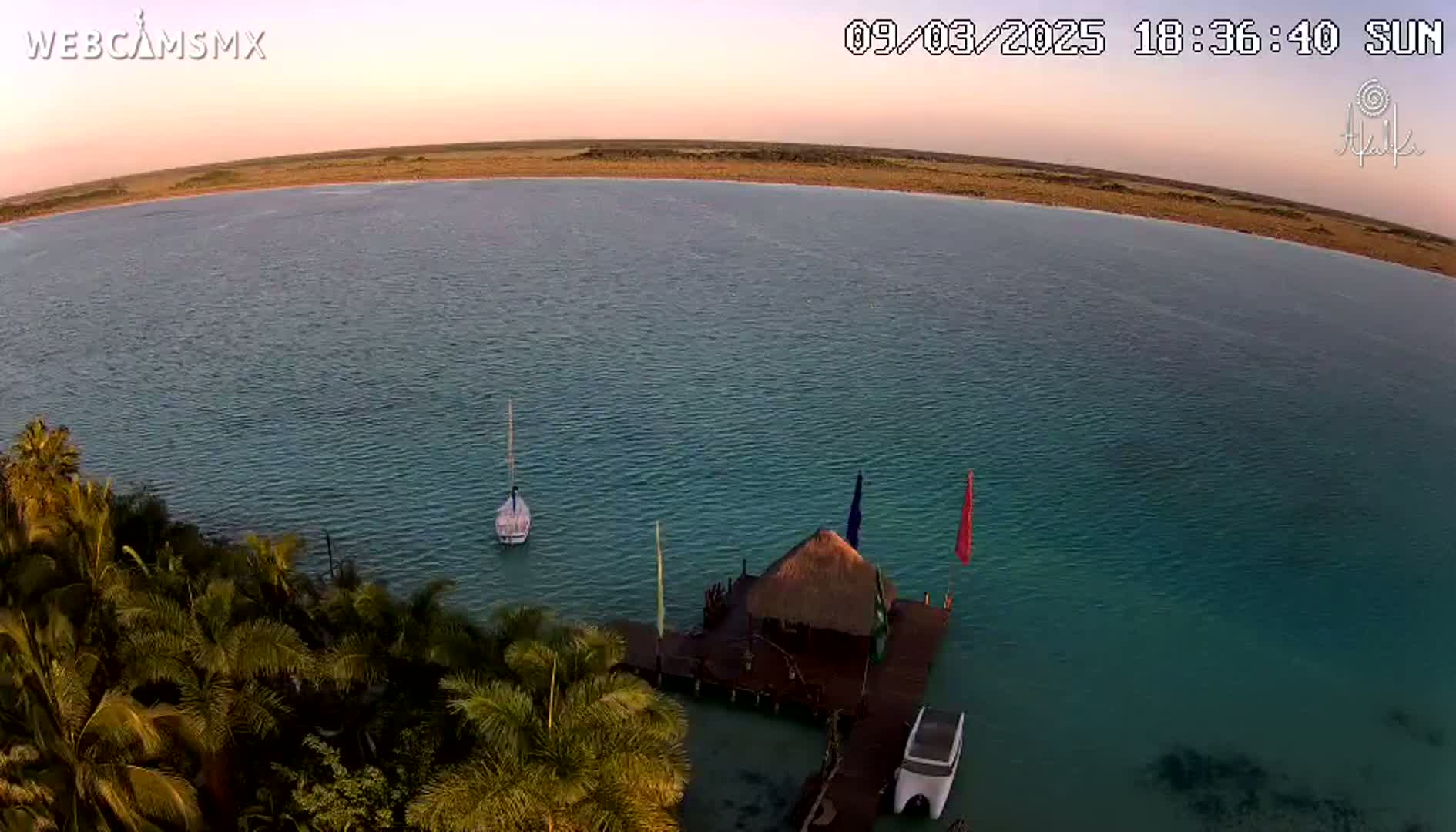 An aerial view shows a calm body of teal water under a sunset sky, with a small sailboat, a thatched-roof structure on a dock, and palm trees along the shoreline.