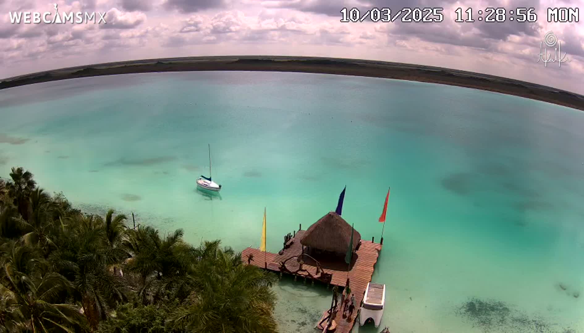 An aerial view shows a sailboat and a thatched-roof structure on a dock in a calm, teal-colored body of water under a partly cloudy sky.