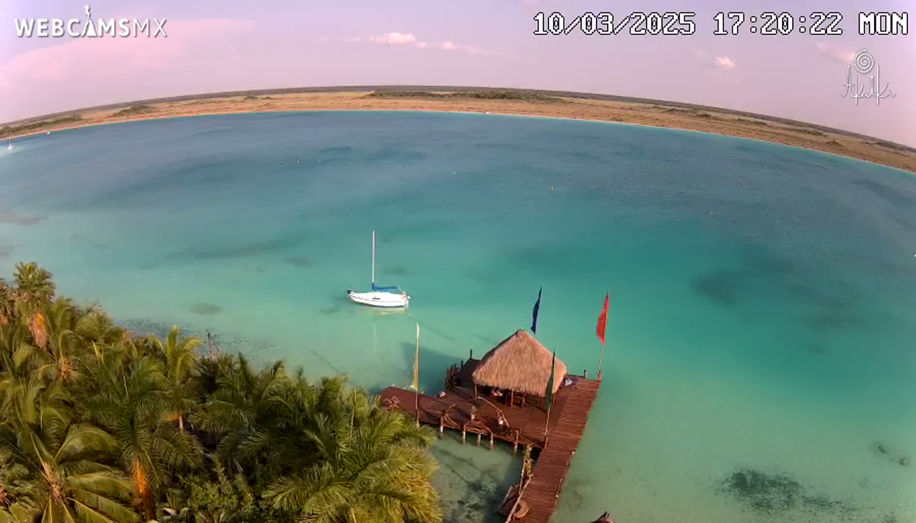An aerial view shows a tranquil, turquoise body of water with a small sailboat and a thatched-roof hut on a pier, surrounded by lush palm trees under a partly cloudy sky.