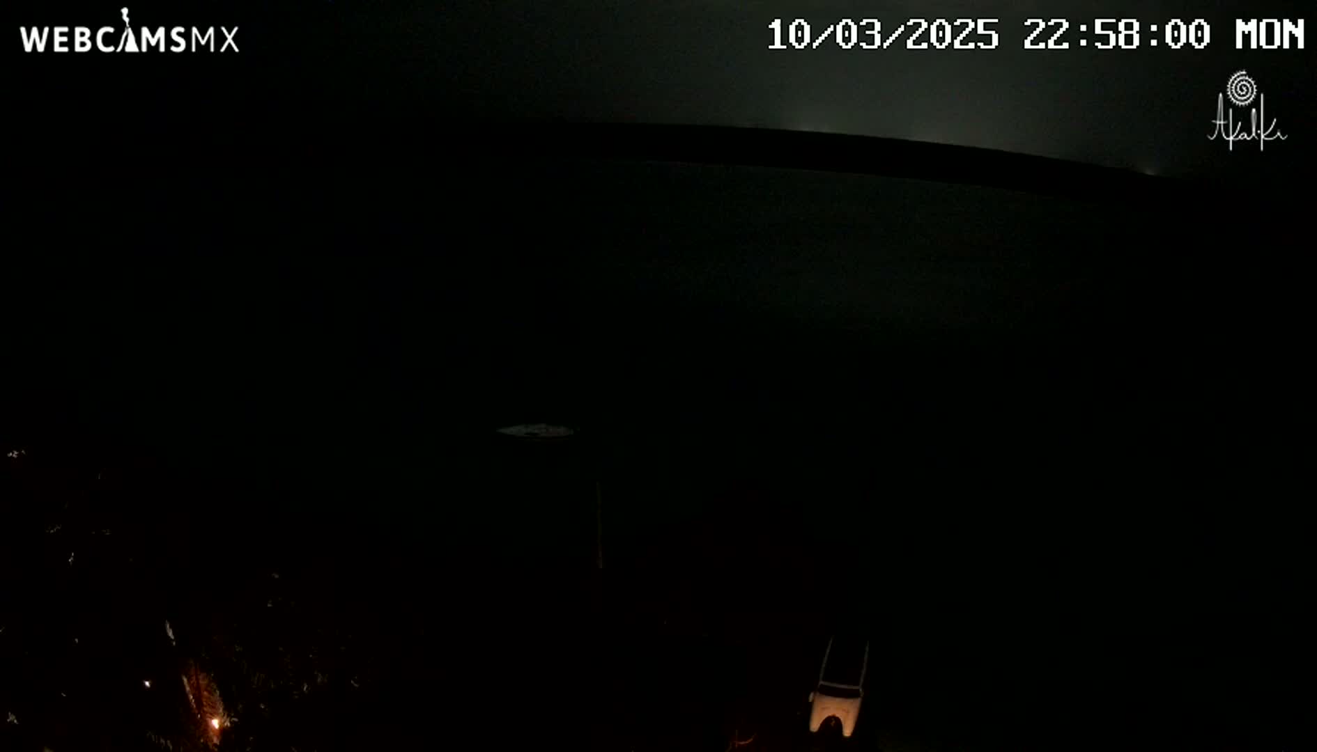 A dark nighttime scene shows a mostly black landscape with a few small lights visible in the bottom left corner and a light-colored object in the bottom right.