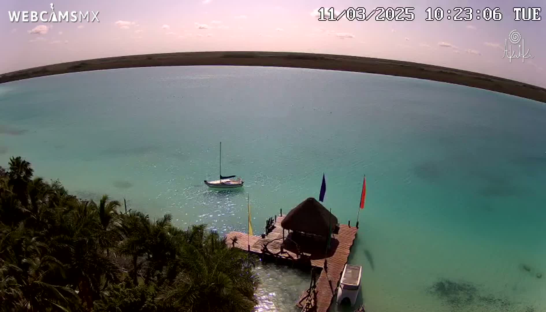 An aerial view shows a sailboat anchored in a calm, teal-colored body of water near a wooden dock with a thatched-roof structure, surrounded by lush green vegetation under a mostly clear sky.