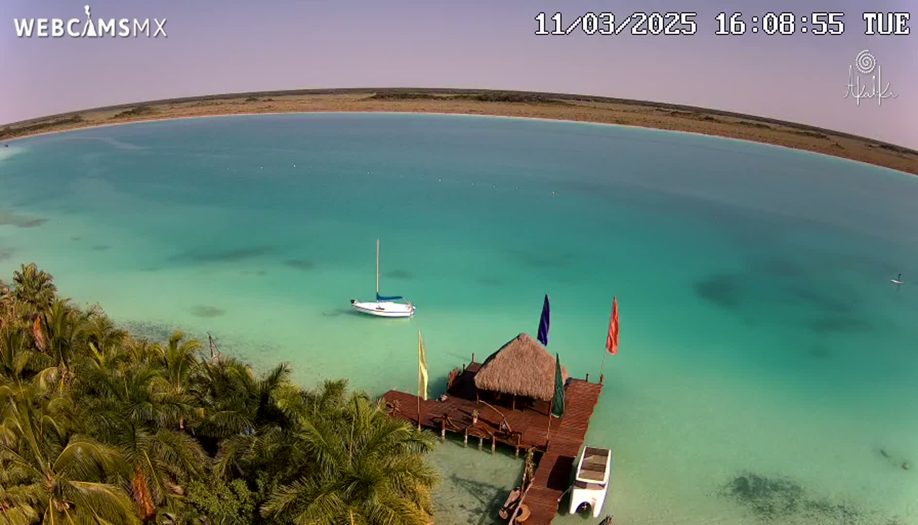 An aerial view shows a tranquil, turquoise body of water with a sailboat, a small thatched-roof hut on a dock, and palm trees lining the shore on a sunny day.