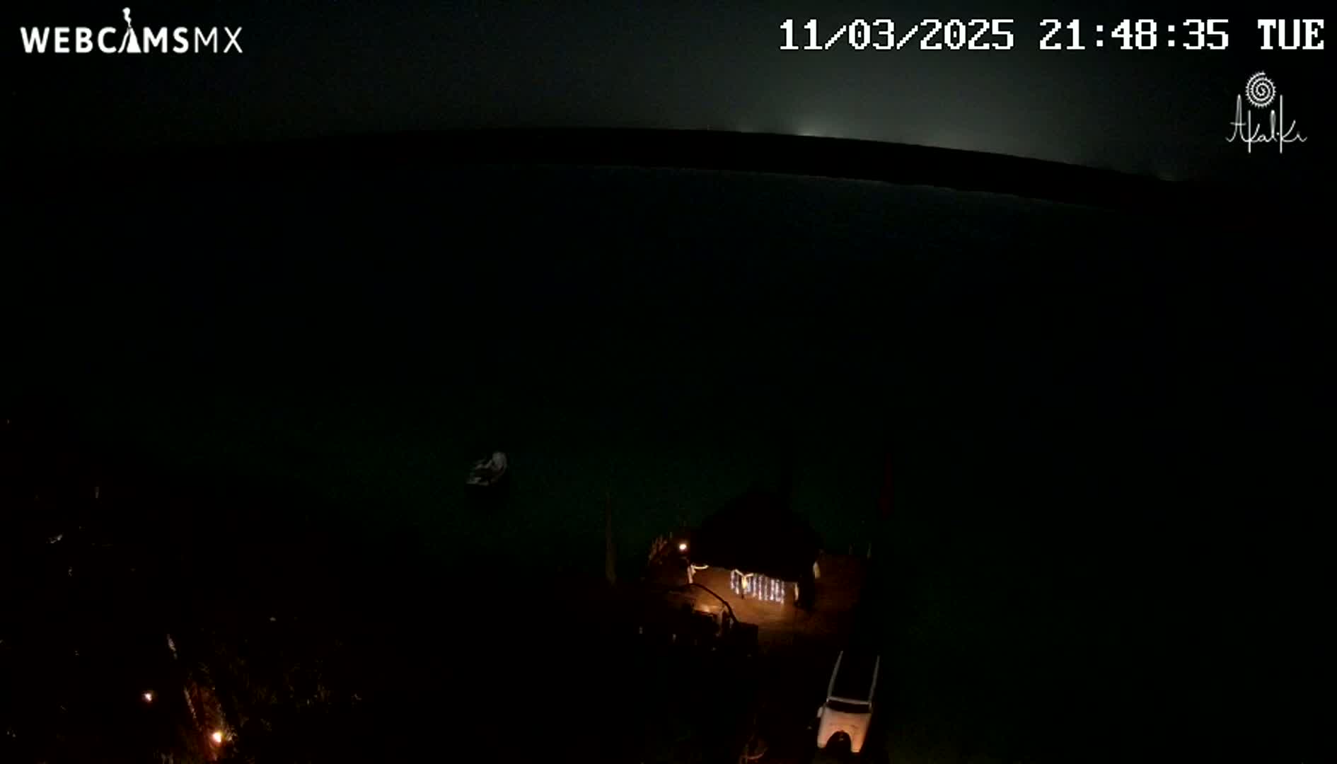 A nighttime, dark view of a body of water shows a small structure with lights on a dock, and a small object floating in the water.
