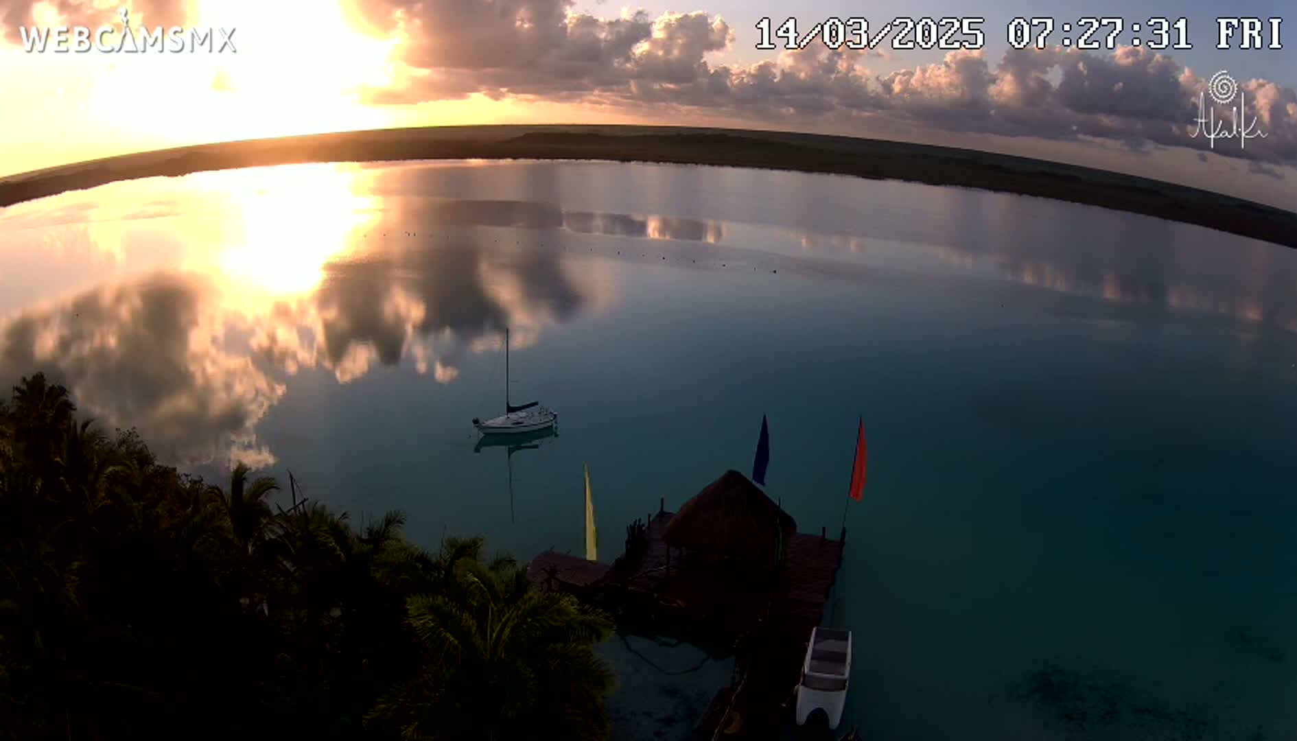 A sunrise casts golden light on a calm body of water, reflecting clouds in its surface, with a sailboat and a small dock with flags visible near a shoreline of tropical vegetation.