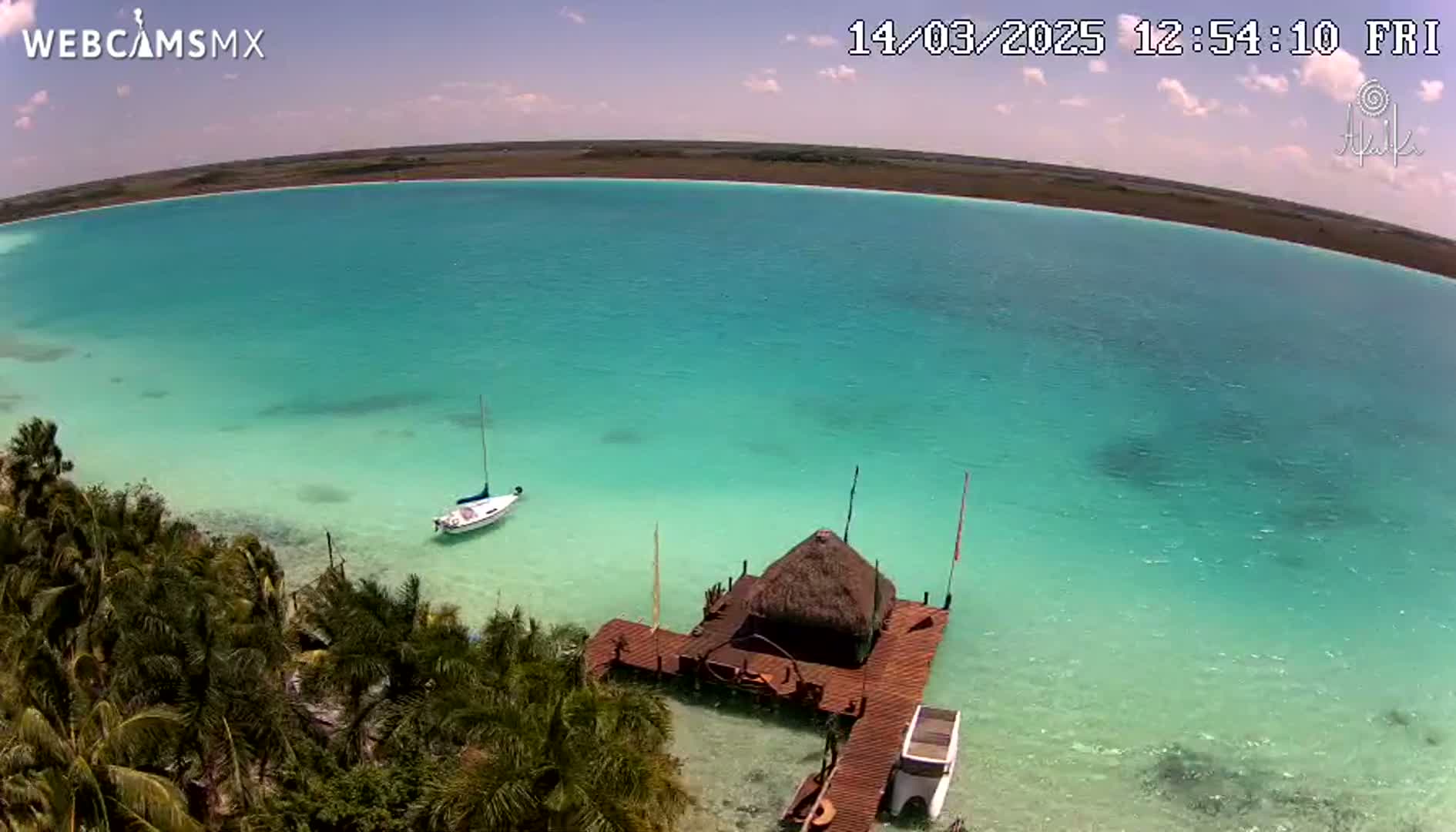 An aerial view shows a tranquil, turquoise body of water with a sailboat and a thatched-roof hut on a wooden dock, under a mostly sunny sky.