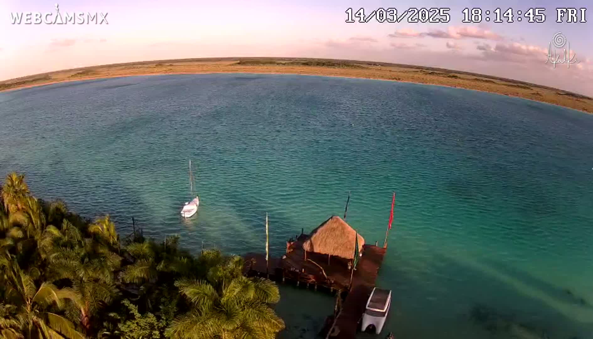 An aerial view shows a calm, teal body of water with a small sailboat and a thatched-roof hut on a dock near a shoreline of lush palm trees under a partly cloudy, dusky sky.
