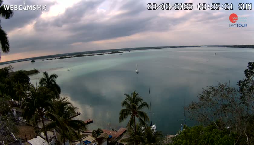 A mostly cloudy day reveals a tranquil body of water with a sailboat in the distance, surrounded by lush tropical vegetation.