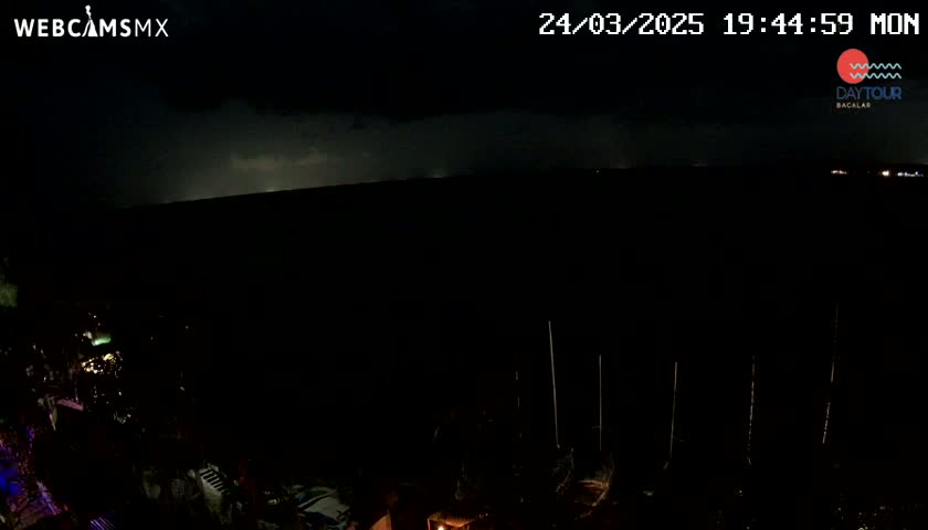 A nighttime view shows distant lightning illuminating a dark, mostly obscured body of water, with some lights visible in the foreground and background.
