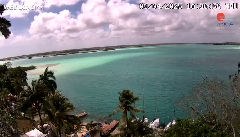 A partly cloudy day reveals a tranquil turquoise lagoon surrounded by lush green vegetation and a few sailboats near the shore.
