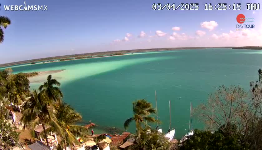 A partly cloudy day reveals a tranquil turquoise lagoon, bordered by lush tropical vegetation and a few sailboats near the shore.
