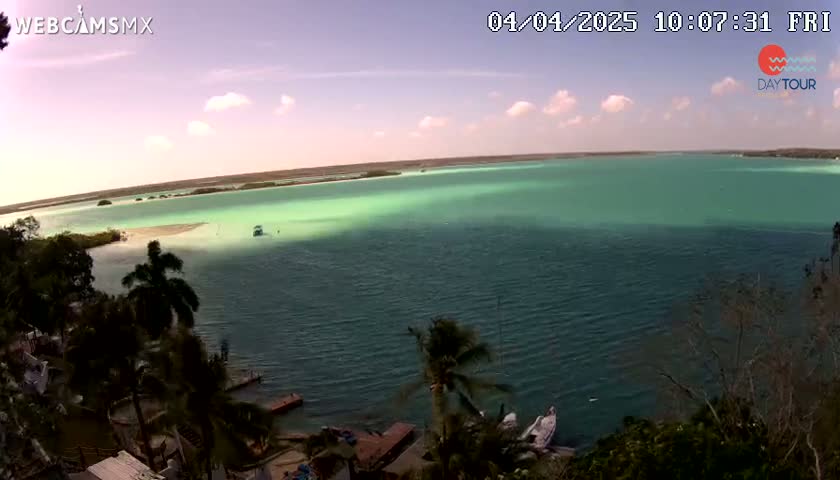 A partly cloudy sky overlooks a calm body of teal-colored water, surrounded by lush green vegetation and palm trees.