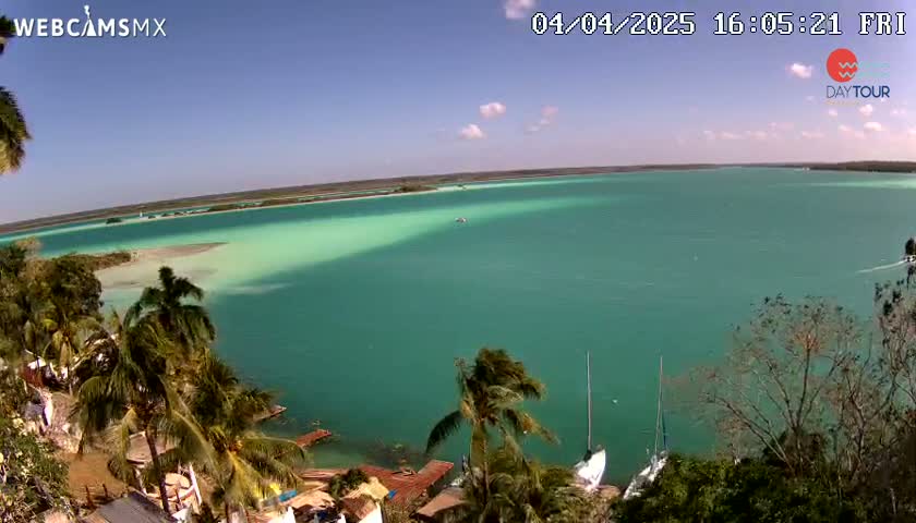 A tranquil turquoise body of water, partially shaded by a shallow, lighter area near the shore, is viewed from an elevated perspective showing palm trees and vegetation on a sunny day with a few clouds.