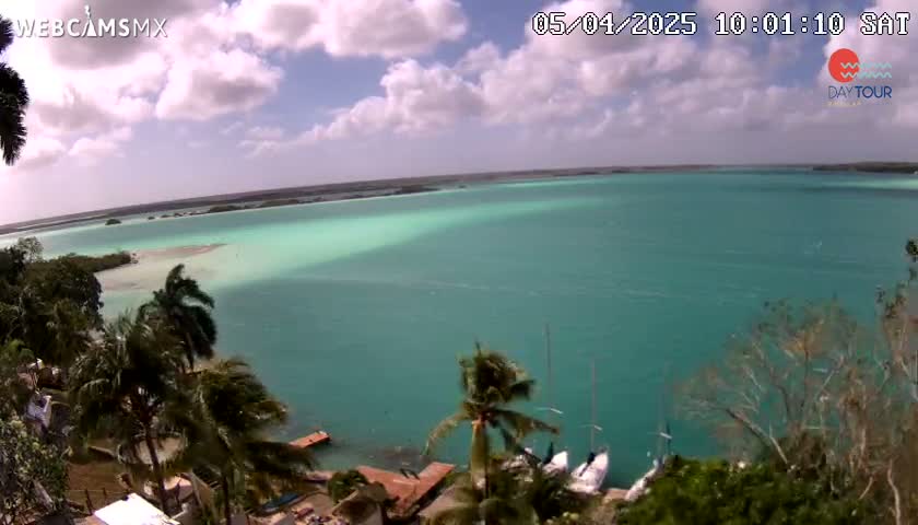 An aerial view shows a partly cloudy day over a teal-colored body of water with several sailboats moored near a shoreline dotted with palm trees and vegetation.