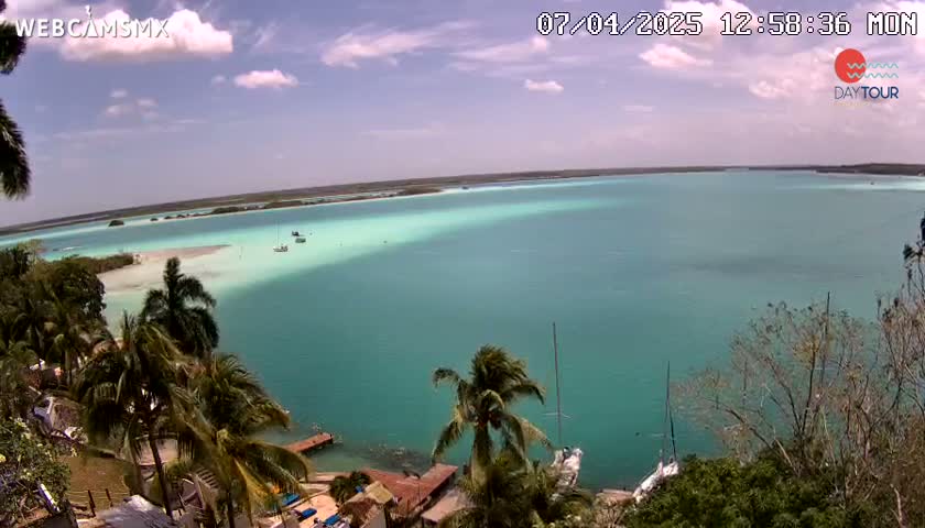 A picturesque, turquoise-colored lagoon is seen from an elevated viewpoint, under a partly cloudy sky, with palm trees and a few boats visible in the distance.