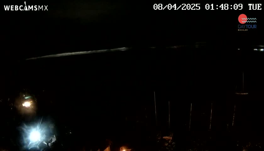 A nighttime, dark scene shows a faint, bright horizon line over a dark body of water, with a vehicle's headlights visible in the lower left corner.
