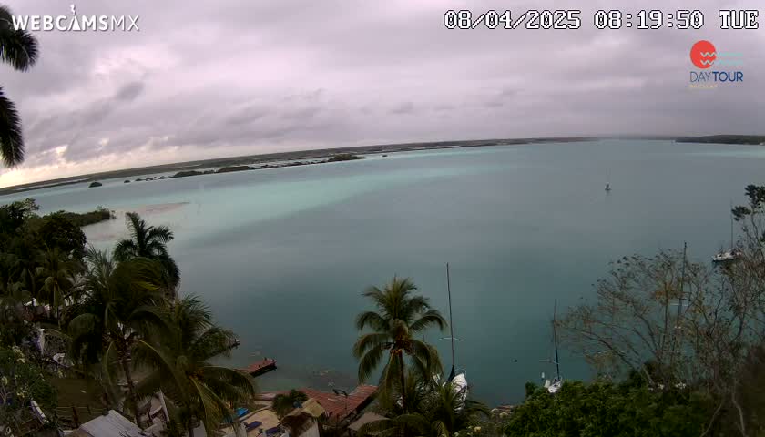 An elevated view of a calm, teal-colored body of water under an overcast sky, surrounded by lush green vegetation and a few sailboats.