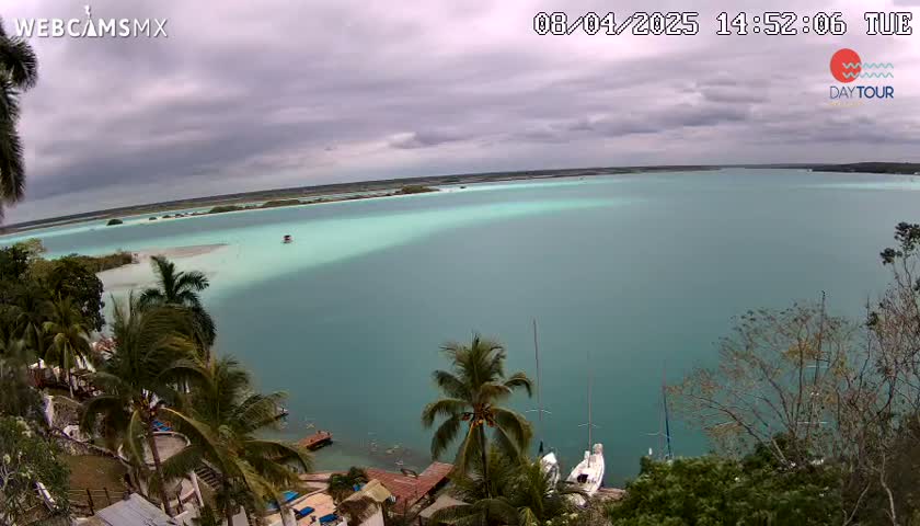 An expansive teal-colored body of water is seen under an overcast sky, bordered by lush green vegetation including palm trees and a shoreline with several small buildings.