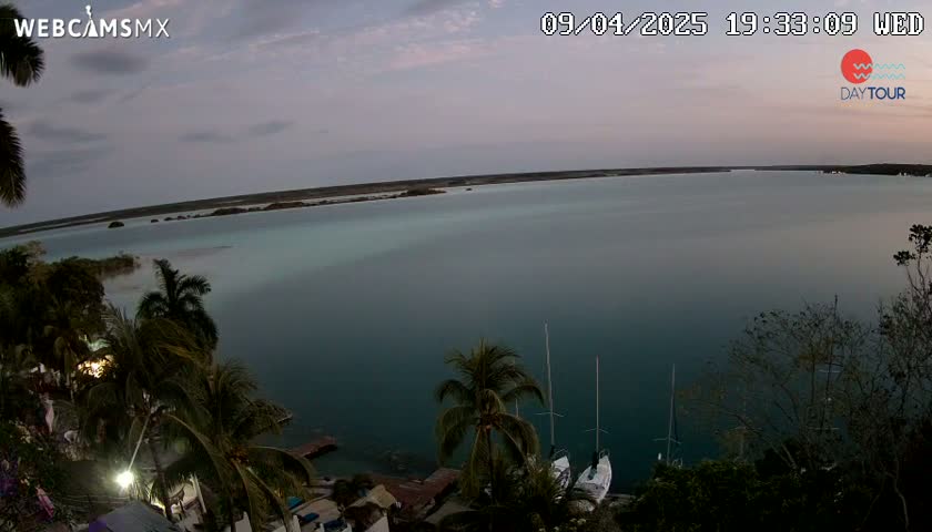 An expansive body of calm, teal water is seen at dusk, surrounded by lush vegetation and two sailboats docked near the shore.
