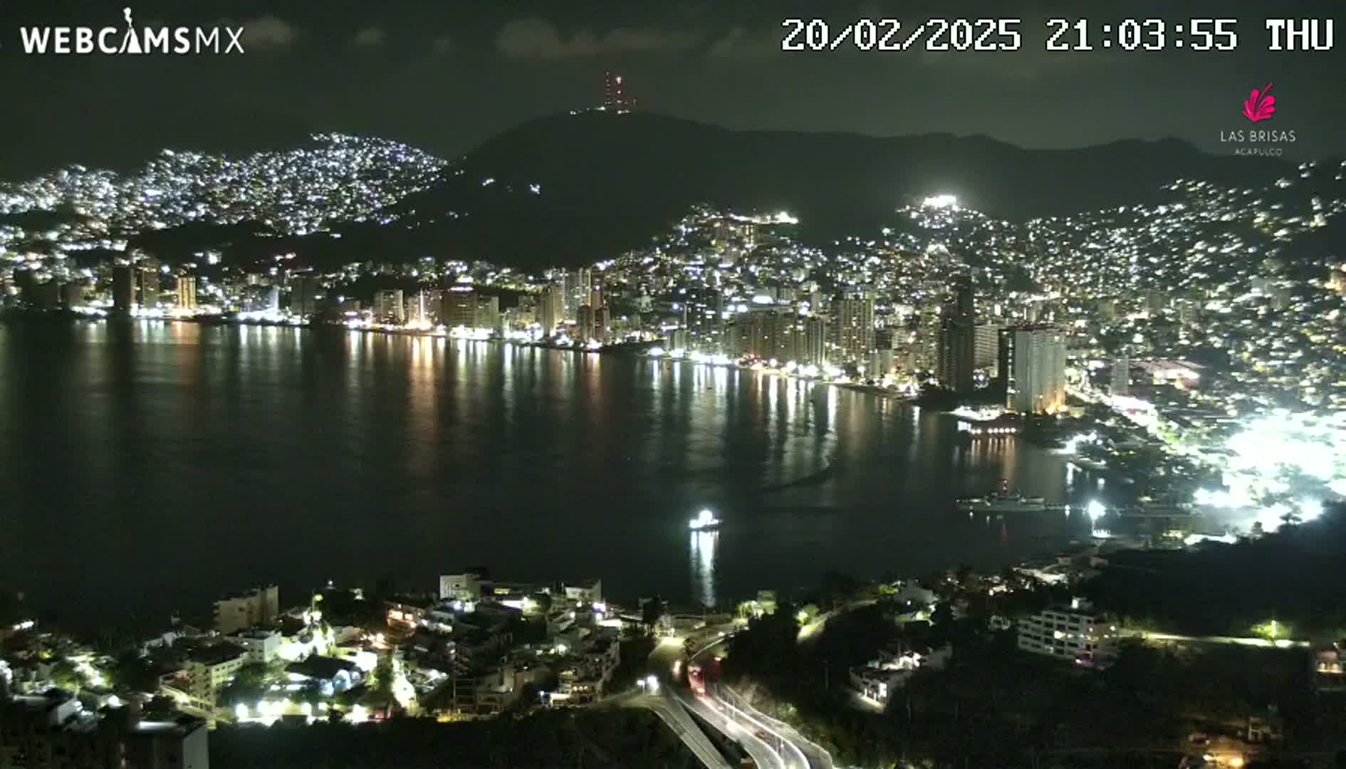 A nighttime view of a city illuminated along a dark bay, nestled against a hill under a clear sky.