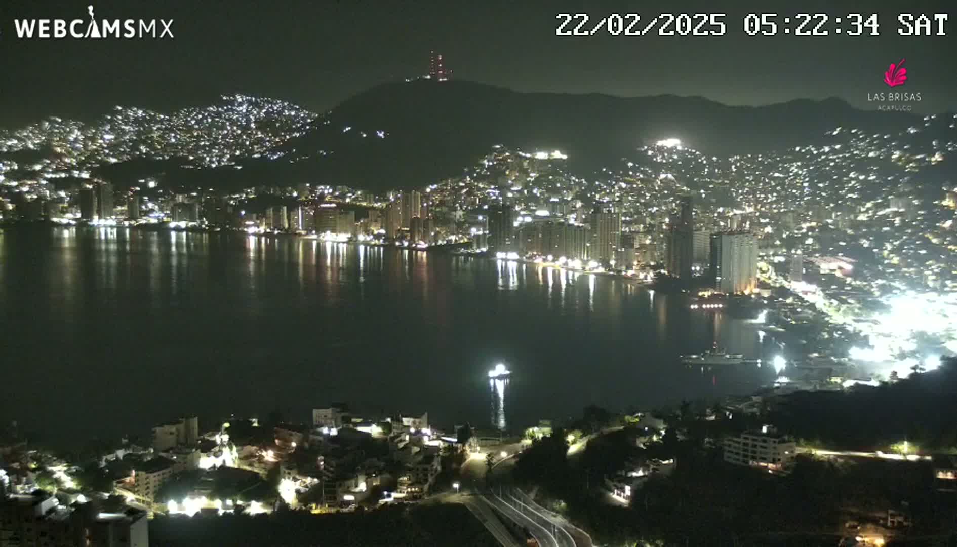 A nighttime, aerial view shows a city sprawling across a hill and along a bay, with its lights reflecting on the dark water.