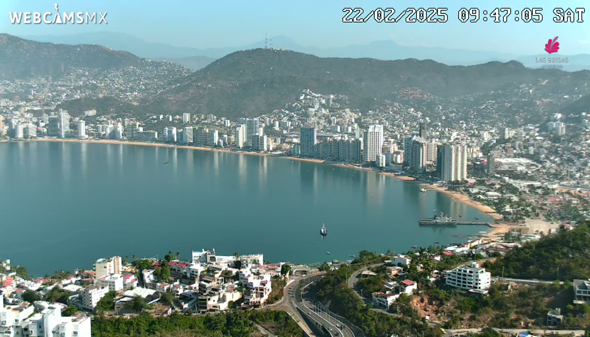 A coastal city with high-rise buildings sprawls along a calm bay under a clear, sunny sky.